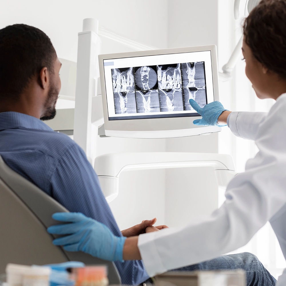 Man seated in a dental chair while the dentist beside him points to dental X-rays displayed on a screen.