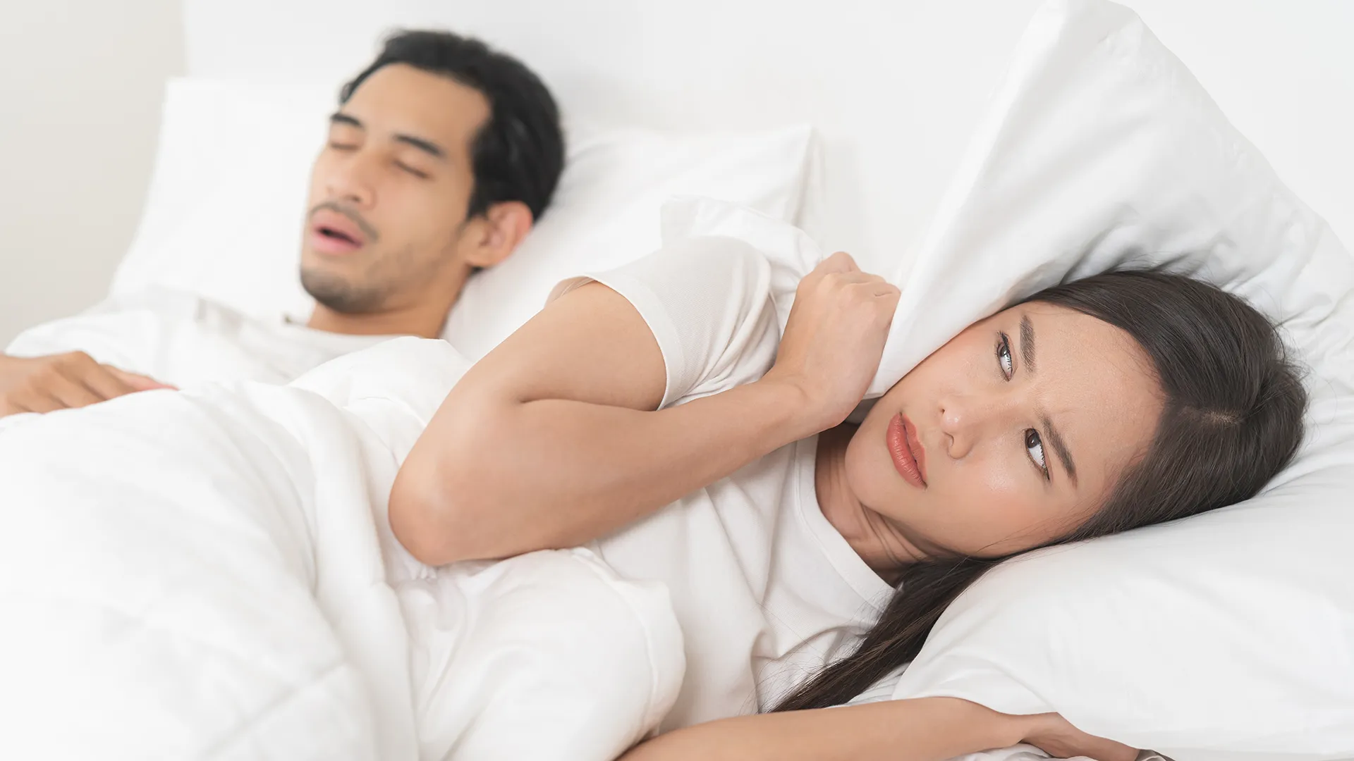 Woman lying in bed with her husband, covering her ears with a pillow due to his snoring.