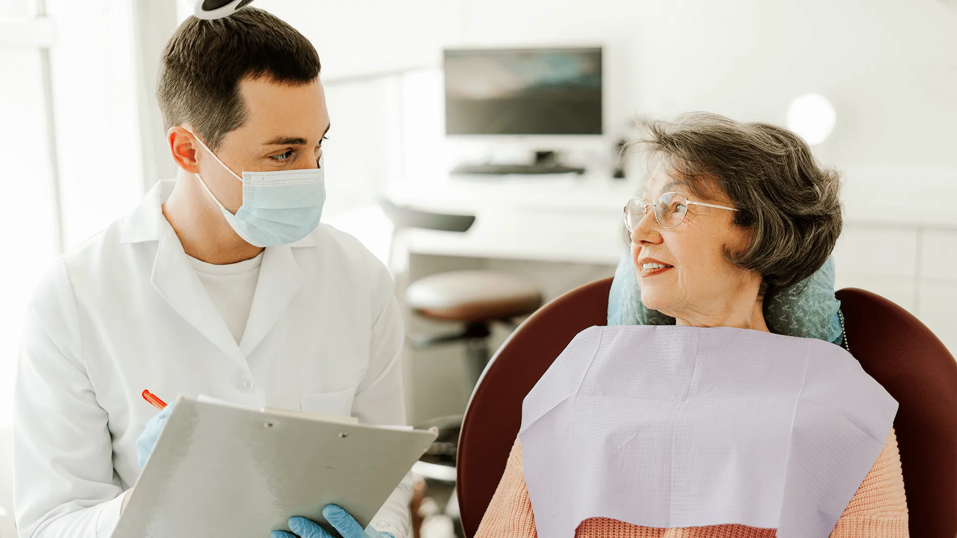 Older woman sitting in a dental chair while the masked dentist takes notes.
