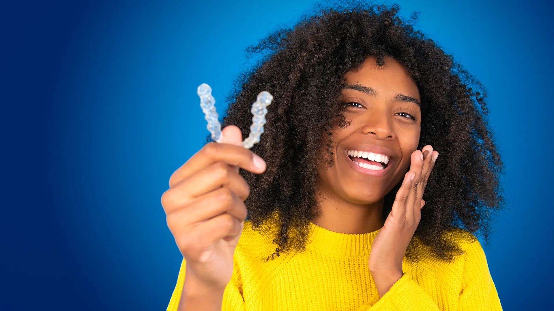 Woman smiling against a blue background while holding clear aligners.
