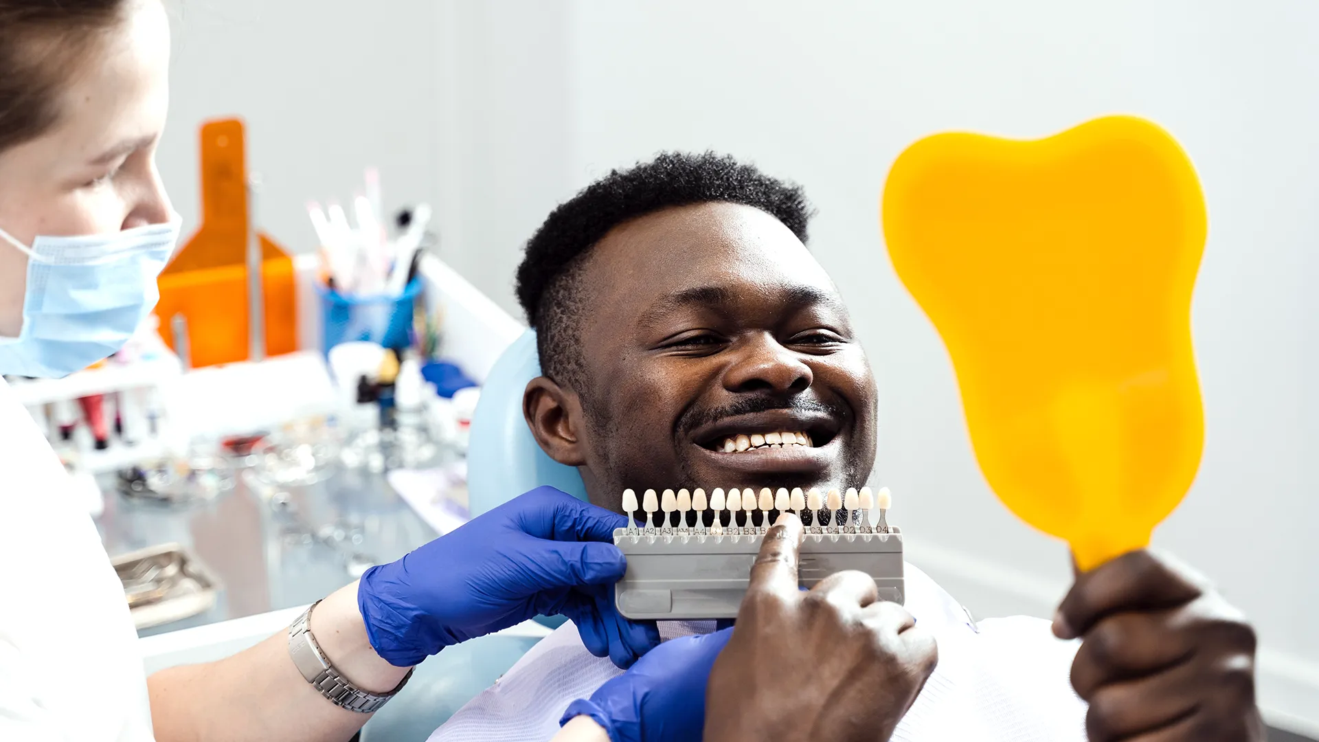 Man sitting in a dental chair holding a mirror and smiling while the dentist positions a set of veneers in front of his teeth.