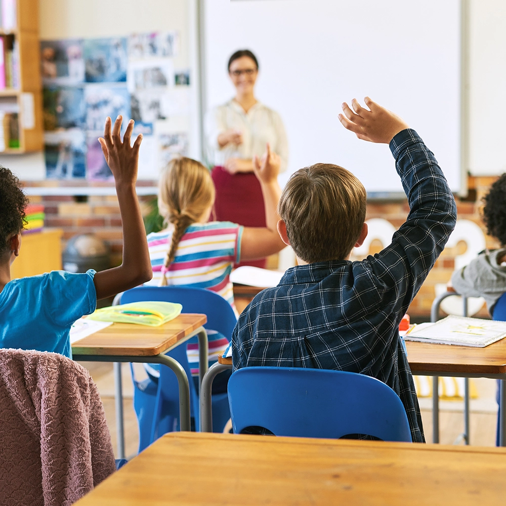 Children sitting in a classroom raising their hands while looking at the teacher.