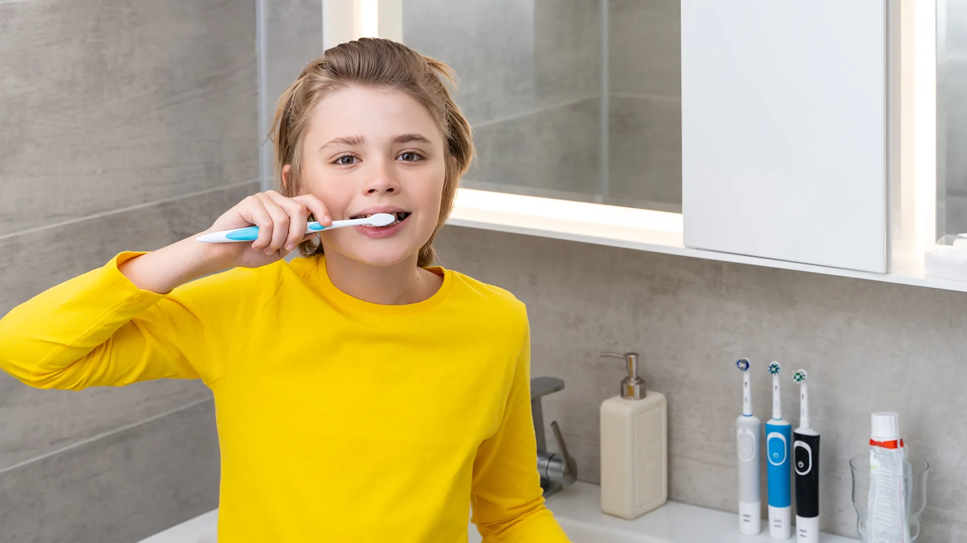 Child wearing a yellow sweater brushing their teeth while looking at the camera.