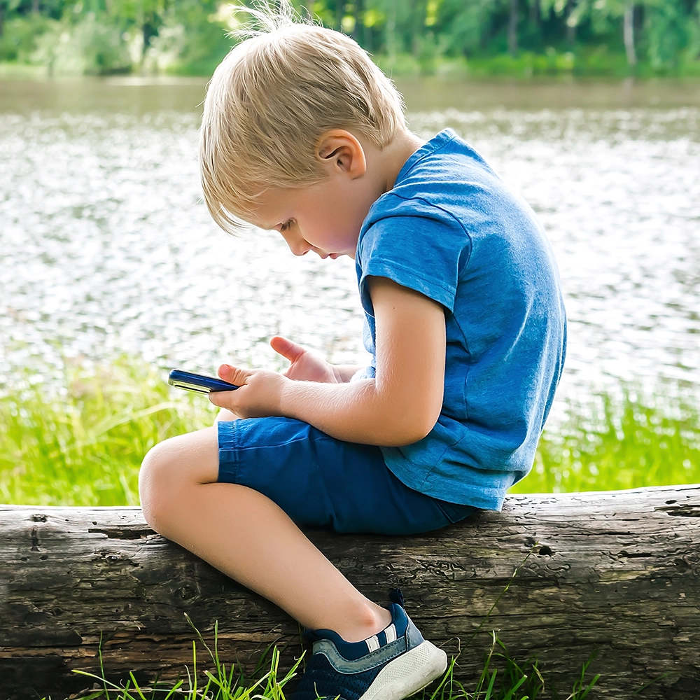 Child sitting on a wooden log watching something on a smartphone.