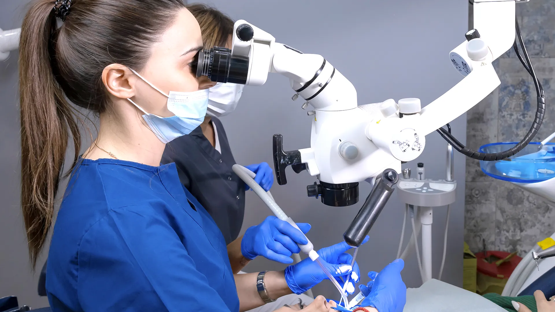 Dentist performing a procedure with dental tools and a microscope while the patient lies on the dental chair.