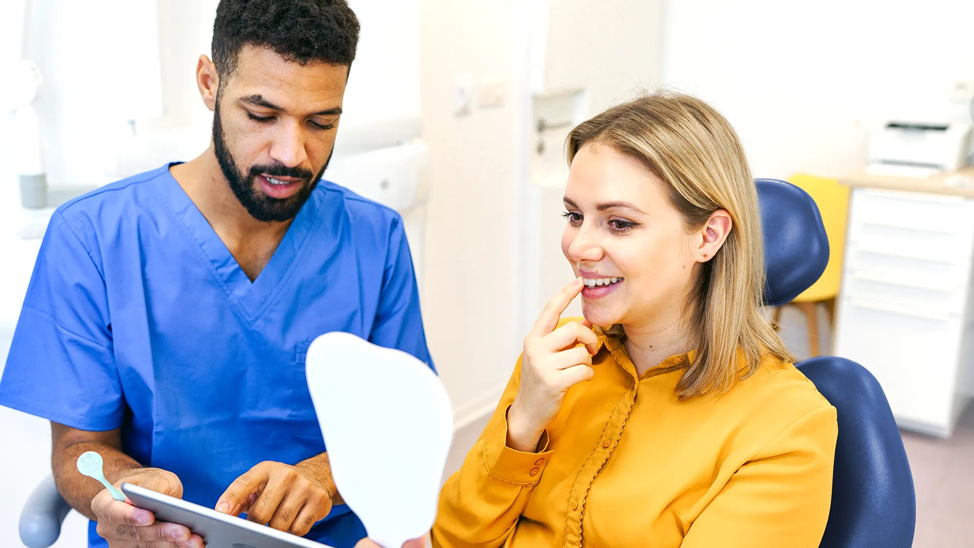 Woman sitting in a dental chair pointing at her tooth in the mirror while the dentist shows her information on a tablet.