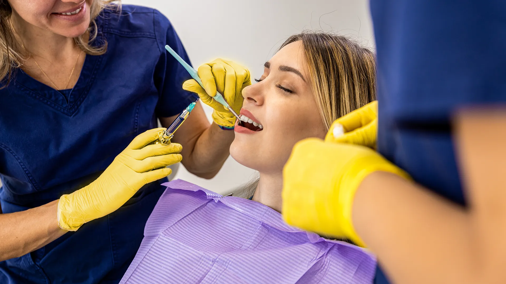 patient lying in a dental chair with mouth open while a dentist holds a syringe.
