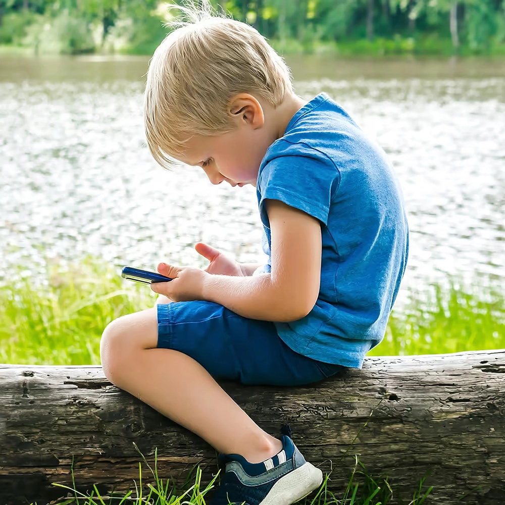 Child sitting on a wooden log watching something on a smartphone.