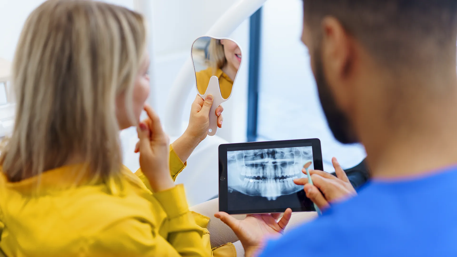 Woman holding a mirror to check her teeth while the dentist beside her reviews dental X-rays on a tablet.