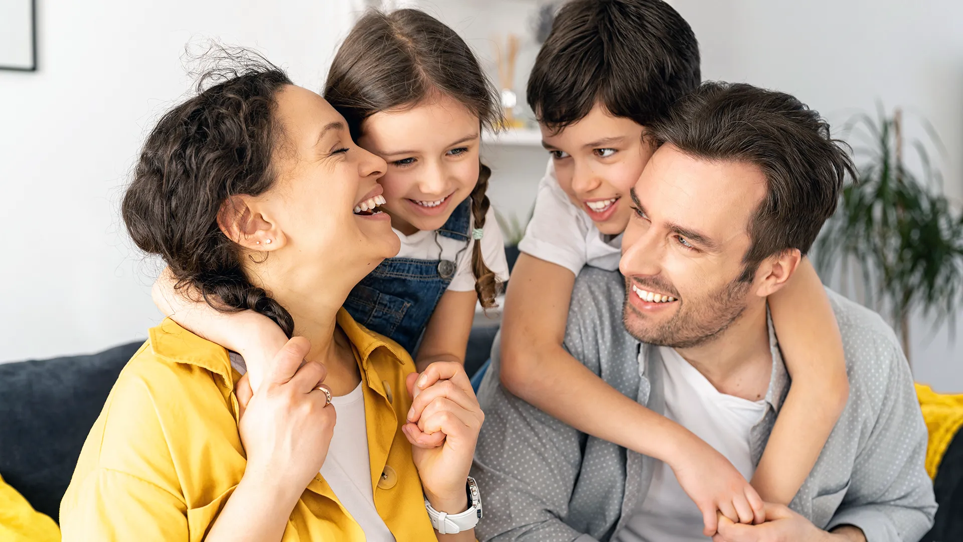 Seated parents smiling at each other with their daughter on the mother’s back and their son on the father’s back.
