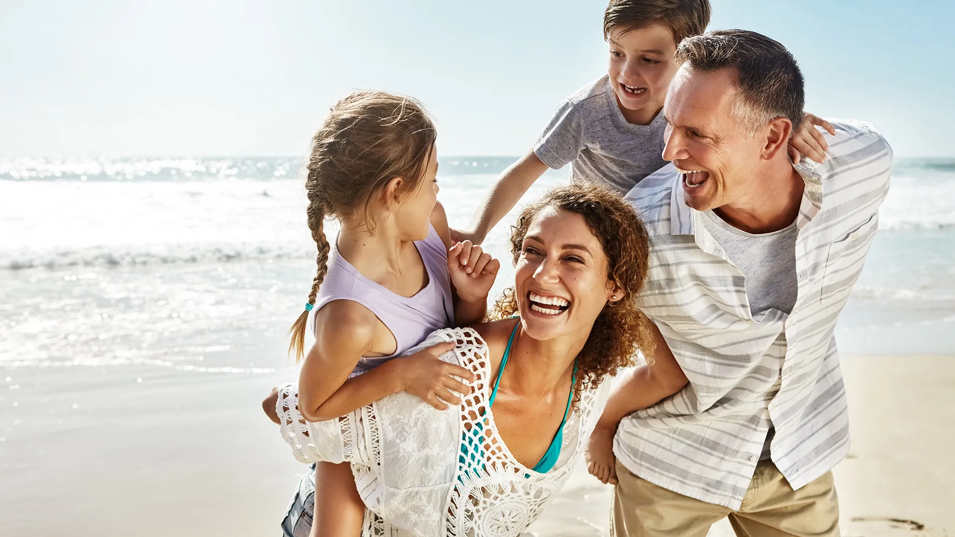 Family at the beach with parents carrying two children—one girl and one boy—on their backs, all laughing.